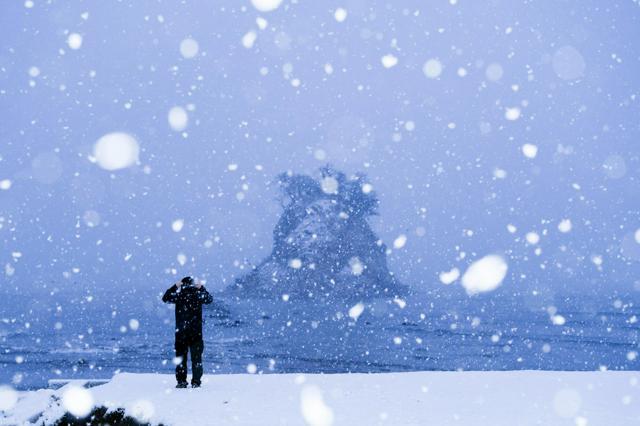 雪が降りしきる中、海岸から見附島を見つめる人