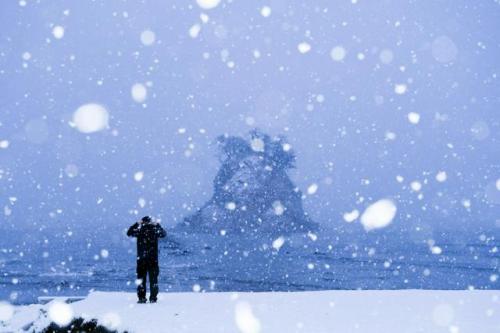雪が降りしきる中、海岸から見附島を見つめる人